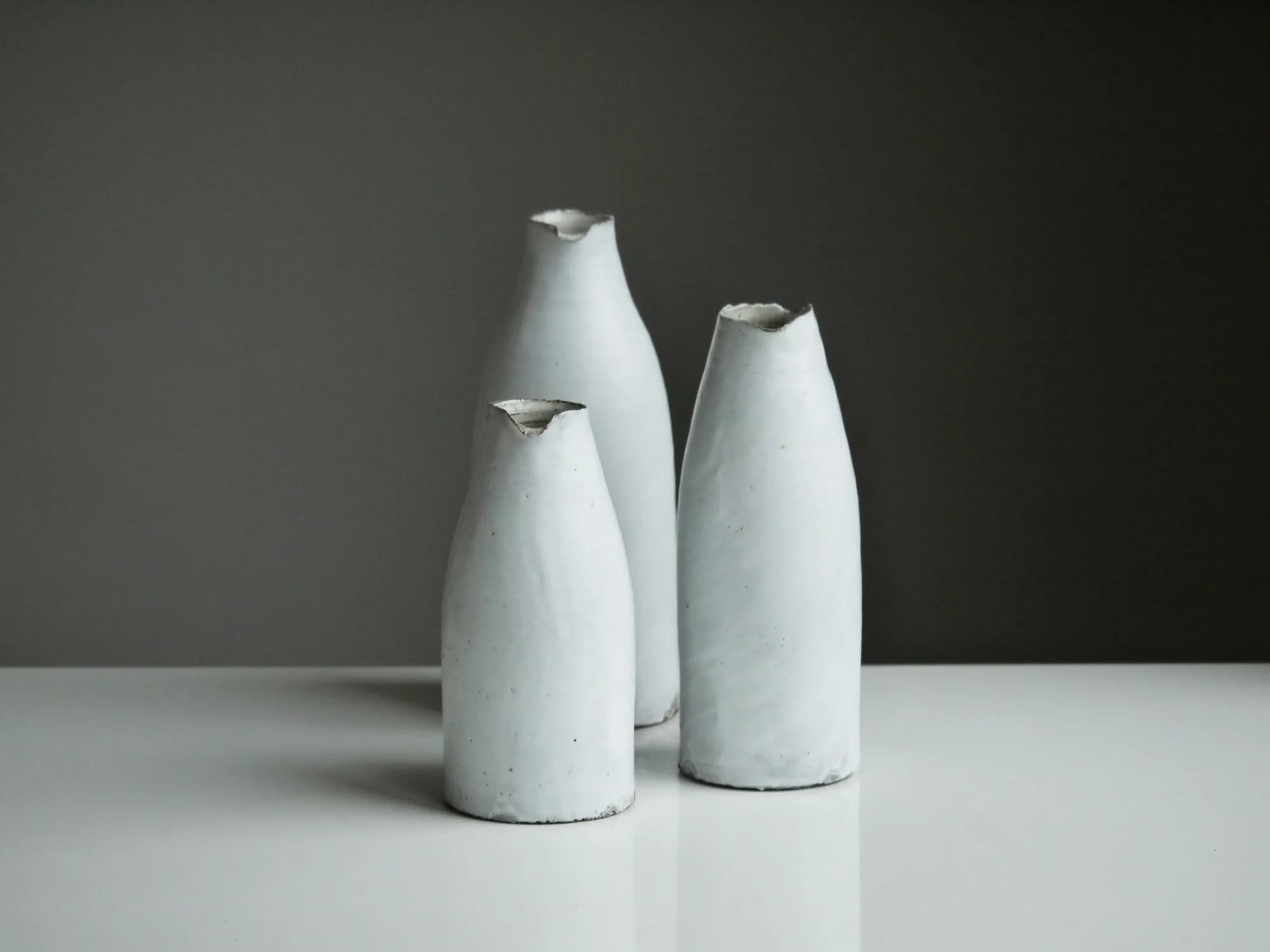Trois vases de céramique claire de Tom Crew, aux silhouettes irrégulières, photographiés en lumière naturelle.