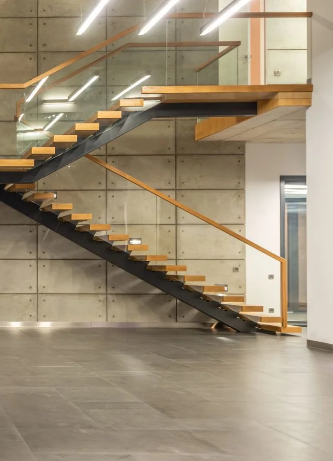 Détail de l’escalier : marches de bois, garde-corps vitré et béton. Recadrage de la vue intérieure.